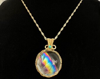 May include: A gold chain necklace with a round pendant featuring a clear crystal with iridescent rainbow colors inside. The pendant is surrounded by a gold wire cage and has a small turquoise stone at the top.