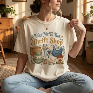 May include: Cream-colored t-shirt with the text "Take Me To The Thrift Shop" in blue and yellow. The graphic features a bread box, bowls, a duck, a chicken, and a mushroom. The shirt is worn by a person.