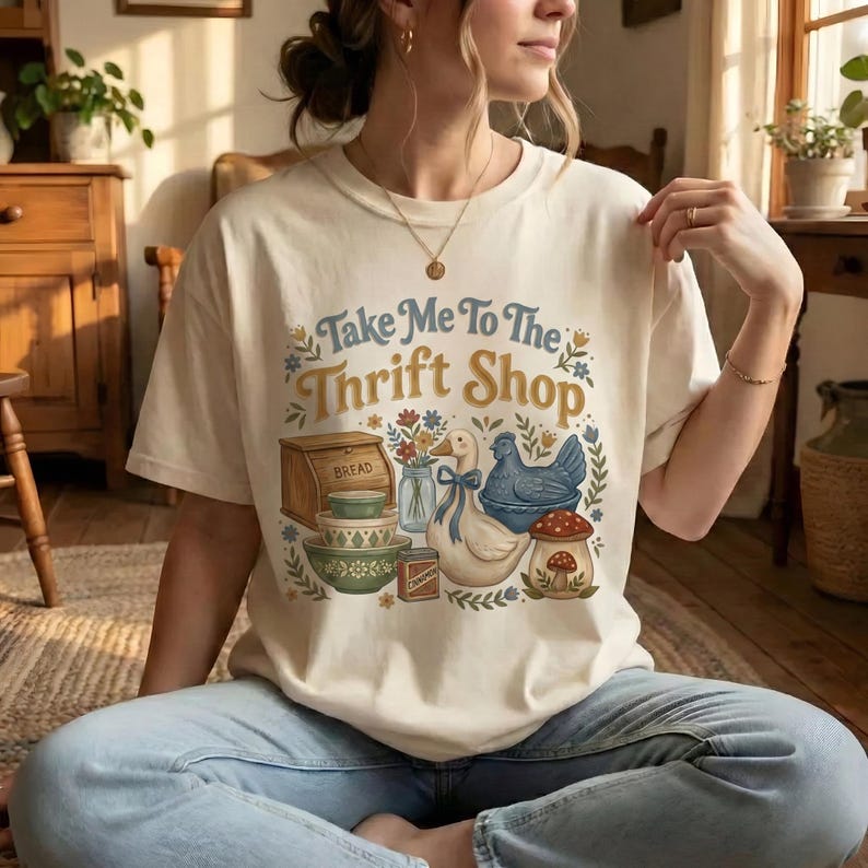 May include: Cream-colored t-shirt with the text "Take Me To The Thrift Shop" in blue and yellow. The graphic features a bread box, bowls, a duck, a chicken, and a mushroom. The shirt is worn by a person.