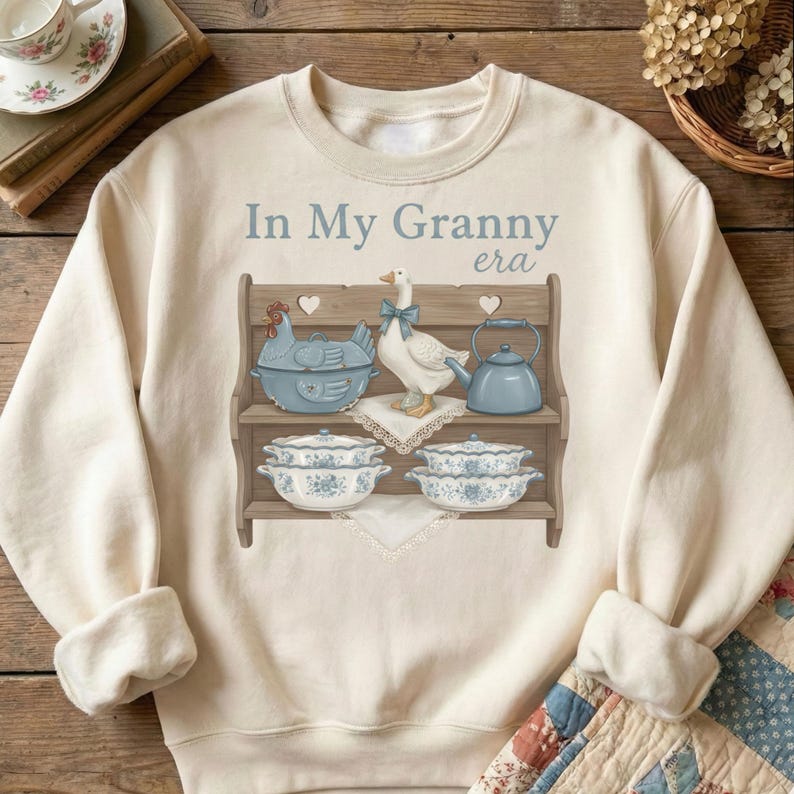 Op de afbeelding: Cr&egrave;mekleurige sweatshirt met de tekst "In My Granny era" boven een afbeelding van een houten plank met blauw en wit keukengerei en een gans met een blauwe strik. Het ontwerp bevat een kipvormige schaal, een theepot en bloemschalen.
