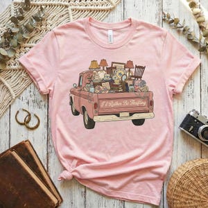 May include: A light pink t-shirt featuring a vintage pink pickup truck filled with various antique items. The truck bed is overflowing with lamps, chairs, dishes, and other vintage finds. The text "I'd Rather Be Thrifting" is printed on the truck.