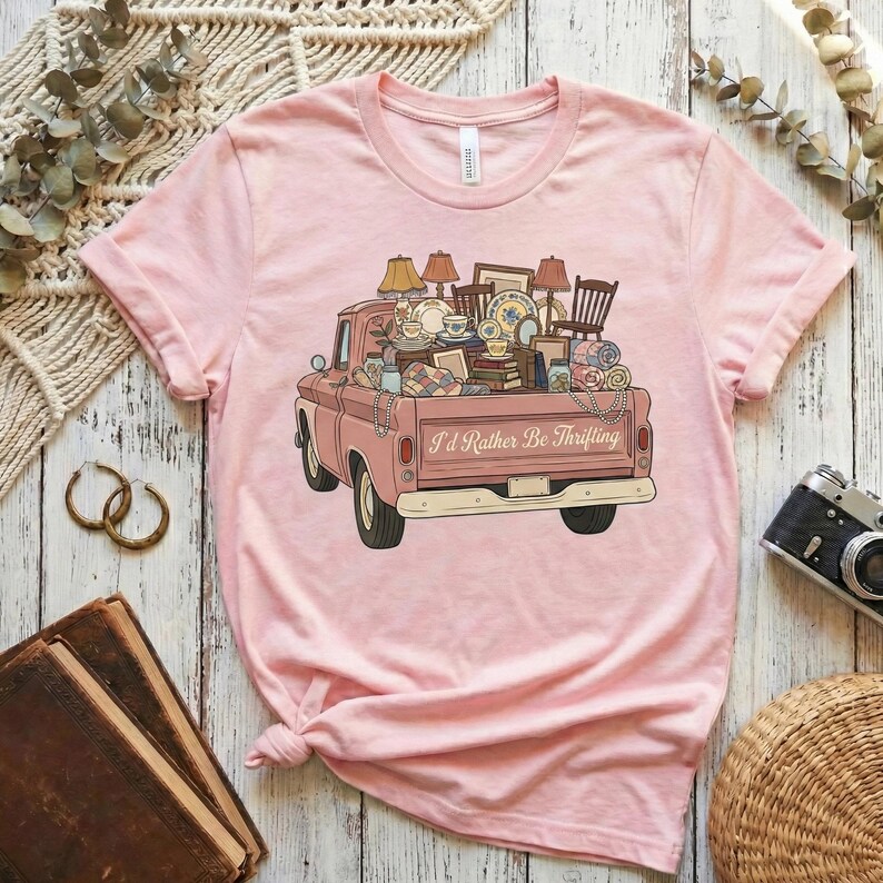 May include: A light pink t-shirt featuring a vintage pink pickup truck filled with various antique items. The truck bed is overflowing with lamps, chairs, dishes, and other vintage finds. The text "I'd Rather Be Thrifting" is printed on the truck.