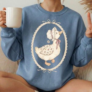 May include: A light blue sweatshirt featuring a vintage-style illustration of a goose wearing a bonnet and a pink bow. The goose is inside an oval frame with a decorative border. The sweatshirt is a solid color and has long sleeves.