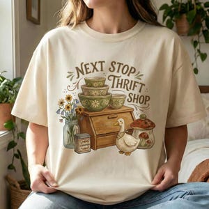 May include: Cream-colored t-shirt with a whimsical graphic. The design features stacked green bowls, a wooden breadbox, a duck, and a mushroom. Text reads "NEXT STOP THRIFT SHOP" in brown lettering. A jar of flowers and a cinnamon tin are also included.
