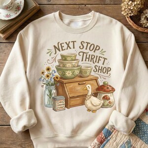May include: Cream-colored sweatshirt with a graphic featuring a wooden breadbox, bowls, a duck, and the text "NEXT STOP THRIFT SHOP." The design includes a jar of flowers, a spice tin, and a mushroom-shaped container. The sweatshirt has ribbed cuffs and a crew neck.