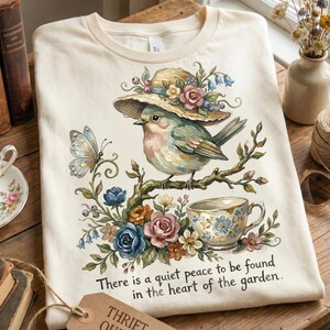Pu&ograve; includere: T-shirt color crema con un'illustrazione ad acquerello di un uccello con un cappello, appollaiato su un ramo con fiori e una tazza da t&egrave;. Il testo recita: "C'&egrave; una quieta pace da trovare nel cuore del giardino."