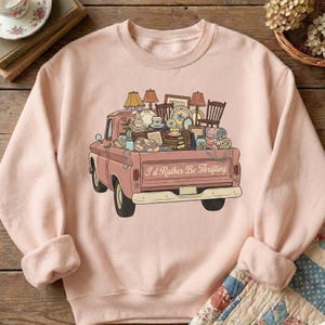 May include: A light pink sweatshirt featuring a vintage pink pickup truck filled with thrifted treasures. The truck bed is overflowing with lamps, chairs, dishes, and other items. The text "I'd Rather Be Thrifting" is printed on the truck.