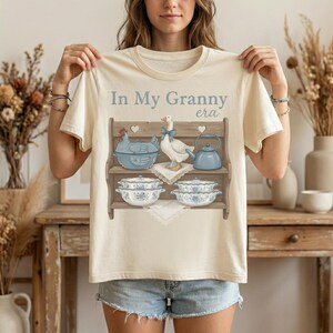 Op de afbeelding: Cr&egrave;mekleurig T-shirt met de tekst "In My Granny era" boven een afbeelding van een plank met blauw en wit keukengerei, waaronder een theepot, kommen en een gans met een blauwe strik.