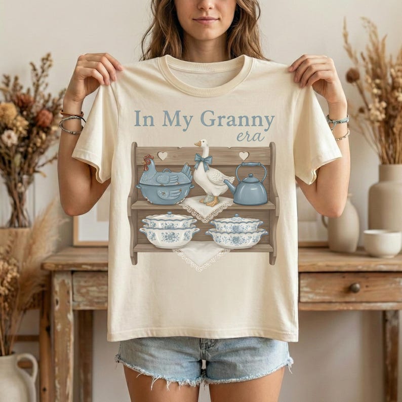 Op de afbeelding: Cr&egrave;mekleurig T-shirt met de tekst "In My Granny era" boven een afbeelding van een plank met blauw en wit keukengerei, waaronder een theepot, kommen en een gans met een blauwe strik.