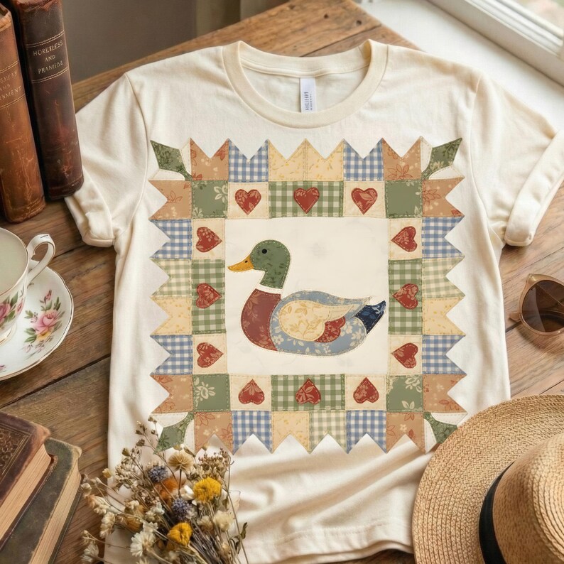 以下が含まれることがあります： クリーム色のTシャツ。パッチワークのデザインで、アヒルのイラストが描かれています。アヒルは緑色の頭、赤い胸、青とベージュの体です。パッチワークには、ハートと緑、ベージュ、青の色合いの様々な模様が含まれています。
