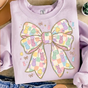 Könnte beinhalten: Ein helllila Sweatshirt mit einem großen Schleifendesign mit goldglitzernden Konturen. Der Bogen ist mit pastellfarbenen Hasen, Küken und Karomustern verziert, ideal für Ostern. Das Sweatshirt ist ein bequemer, lässiger Stil.