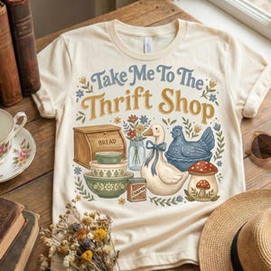 May include: Cream-colored t-shirt with the text "Take Me To The Thrift Shop" in blue and gold. The graphic includes a bread box, bowls, a duck with a blue bow, a chicken, and a mushroom. The design has a vintage, cottagecore aesthetic.
