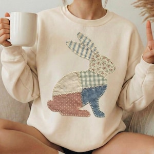 May include: Cream-colored sweatshirt featuring a patchwork bunny design. The bunny is composed of various fabric patterns, including floral, striped, and polka dot. The sweatshirt has long sleeves and a crew neck.