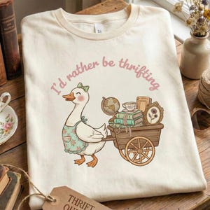 以下が含まれることがあります： ピンク色の文字で「I'd rather be thrifting」と書かれたクリーム色のTシャツ。エプロンを着た漫画のガチョウが、本、地球儀、ティーカップでいっぱいの木製カートを引っ張っています。
