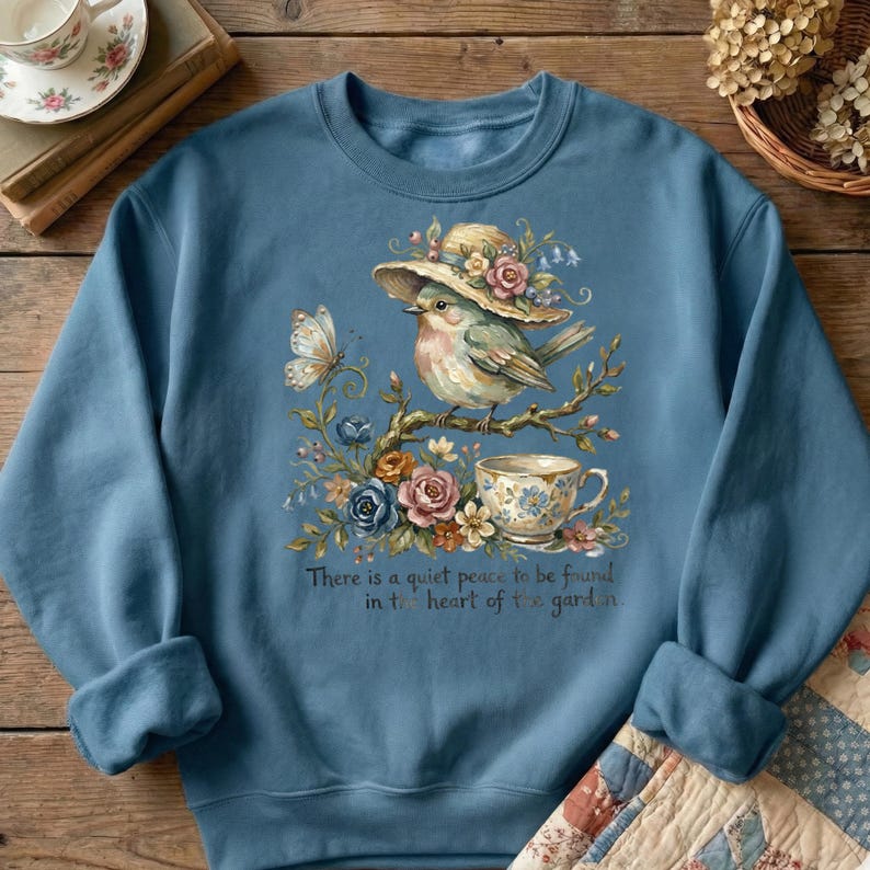Pu&ograve; includere: Felpa blu con un disegno ad acquerello di un uccello con un cappello, appollaiato su un ramo con fiori e una tazza da t&egrave;. Una farfalla e il testo: "C'&egrave; una quieta pace da trovare nel cuore del giardino."