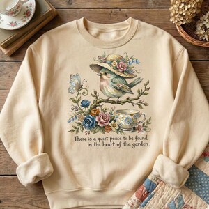 Pu&ograve; includere: Felpa color crema con un'illustrazione dettagliata di un uccello con un cappello, appollaiato su un ramo con fiori e una tazza da t&egrave;. Sono inclusi anche una farfalla e il testo "There is a quiet peace to be found in the heart of the garden".