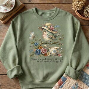 Pu&ograve; includere: Felpa verde salvia con un'illustrazione ad acquerello di un uccello con un cappello, appollaiato su un ramo con fiori e una tazza da t&egrave;. Il testo "There is a quiet peace to be found in the heart of the garden" &egrave; sotto.