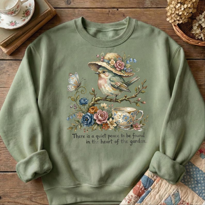 Pu&ograve; includere: Felpa verde salvia con un'illustrazione ad acquerello di un uccello con un cappello, appollaiato su un ramo con fiori e una tazza da t&egrave;. Il testo "There is a quiet peace to be found in the heart of the garden" &egrave; sotto.