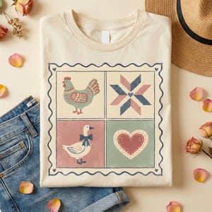 May include: Cream-colored t-shirt featuring a square design with four panels. The panels depict a chicken, a geometric star, a goose with a bow, and a heart. The design is framed with a wavy border and small hearts.