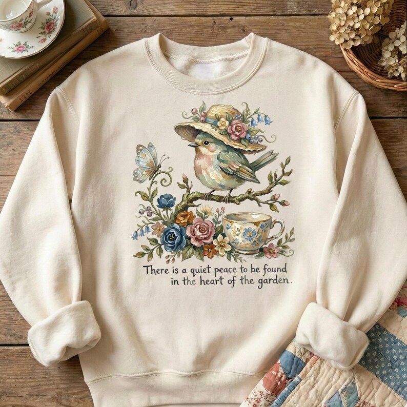 Pu&ograve; includere: Felpa color crema con un'illustrazione ad acquerello di un uccello con un cappello, appollaiato su un ramo fiorito. Sono raffigurati anche una farfalla e una tazza da t&egrave;. Il testo recita: "C'&egrave; una quieta pace da trovare nel cuore del giardino."
