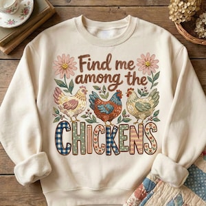 May include: Cream-colored sweatshirt with the text "Find me among the CHICKENS." The graphic features three chickens in shades of brown, blue, and yellow, with floral accents. The word "CHICKENS" is in a decorative font.