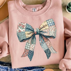 Könnte beinhalten: Ein hellrosa Sweatshirt mit einem großen, farbenfrohen Schleifendesign. Die Schleife besteht aus Patchwork-Stoff mit Blumen-, Vichy- und Streifenmustern in Blau-, Rosa- und Grüntönen. Das Sweatshirt hat einen Rundhalsausschnitt.