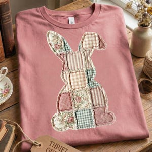 Puede incluir: Una camiseta de color rosa polvoriento con un dise&ntilde;o de conejito de patchwork. El conejito est&aacute; hecho de varios patrones de tela, incluyendo estampados florales, a rayas y a cuadros. La camiseta est&aacute; doblada sobre una superficie de madera.