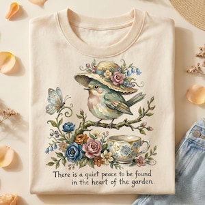Pu&ograve; includere: T-shirt color crema con un'illustrazione ad acquerello di un uccello con un cappello floreale, appollaiato su un ramo con una tazza da t&egrave;. Una farfalla e accenti floreali circondano l'uccello. Il testo recita: "There is a quiet peace to be found in the heart of the garden."