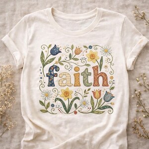 May include: Cream-coloured t-shirt with the word "faith" in colourful, patchwork-style letters. The word is surrounded by a floral design with daisies, tulips, and other flowers. The shirt has short sleeves and a crew neck.