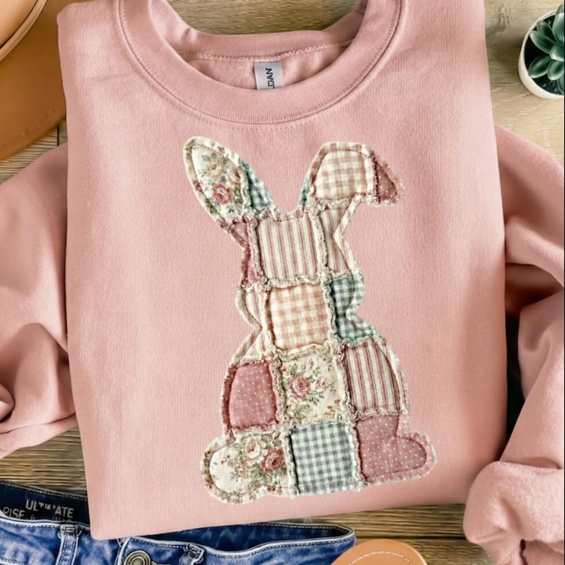 Puede incluir: Una sudadera rosa claro con un dise&ntilde;o de conejo de patchwork. El conejo est&aacute; hecho de varias telas, incluyendo patrones florales, a rayas y a cuadros en tonos de rosa, crema y verde. La sudadera tiene cuello redondo y mangas largas.