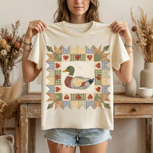 以下が含まれることがあります： パッチワークのアヒルのデザインが施されたクリーム色のTシャツ。アヒルは緑色の頭、黄色のくちばし、赤、青、ベージュのパッチが付いた体を持っています。デザインは、ハートや様々な模様のパッチワークのボーダーで縁取られています。