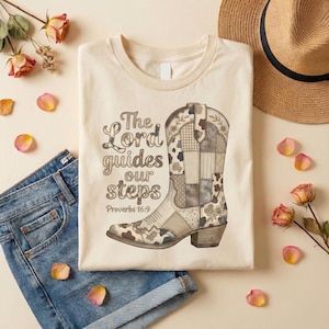 May include: Cream-colored t-shirt featuring a detailed illustration of a patchwork cowboy boot. The boot design incorporates various patterns and textures in shades of brown and gray. The text "The Lord guides our steps Proverbs 16:9" is printed above the boot.