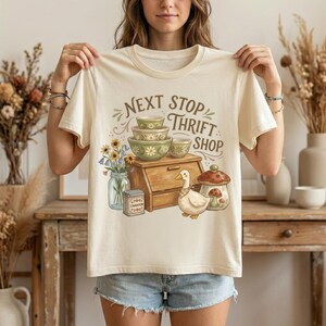 May include: Cream-colored t-shirt with a whimsical illustration. The design features stacked bowls, a wooden cabinet, a duck, and a mushroom. Text reads "NEXT STOP THRIFT SHOP" in a vintage style. A jar of flowers and a spice tin are also included.