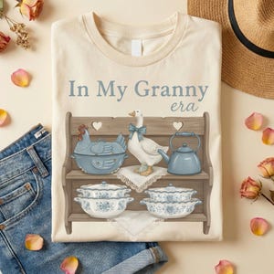 Op de afbeelding: Cr&egrave;mekleurig T-shirt met een afbeelding van een houten plank met een kipvormige ovenschaal, een gans met een blauwe strik, een theepot en bloemige schalen. De tekst "In My Granny era" staat boven de plank.