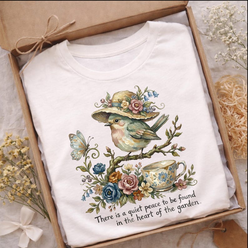 Pu&ograve; includere: T-shirt bianca con un'illustrazione ad acquerello di un uccello con un cappello, appollaiato su un ramo con fiori e una tazza da t&egrave;. Il testo recita: "C'&egrave; una quieta pace da trovare nel cuore del giardino". Presentata in una confezione regalo.