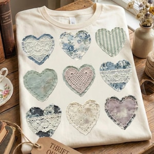 May include: Cream-colored t-shirt with nine heart-shaped patches. The hearts feature various fabrics, including lace, floral prints, and crochet, in shades of blue, pink, and white. The shirt has a classic crew neck.