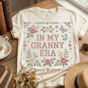 Pode incluir: Camiseta creme com o texto rosa "IN MY GRANNY ERA", rodeada por um design floral com rosas cor-de-rosa, flores azuis e uma borboleta. A camiseta est&aacute; em uma superf&iacute;cie de madeira.