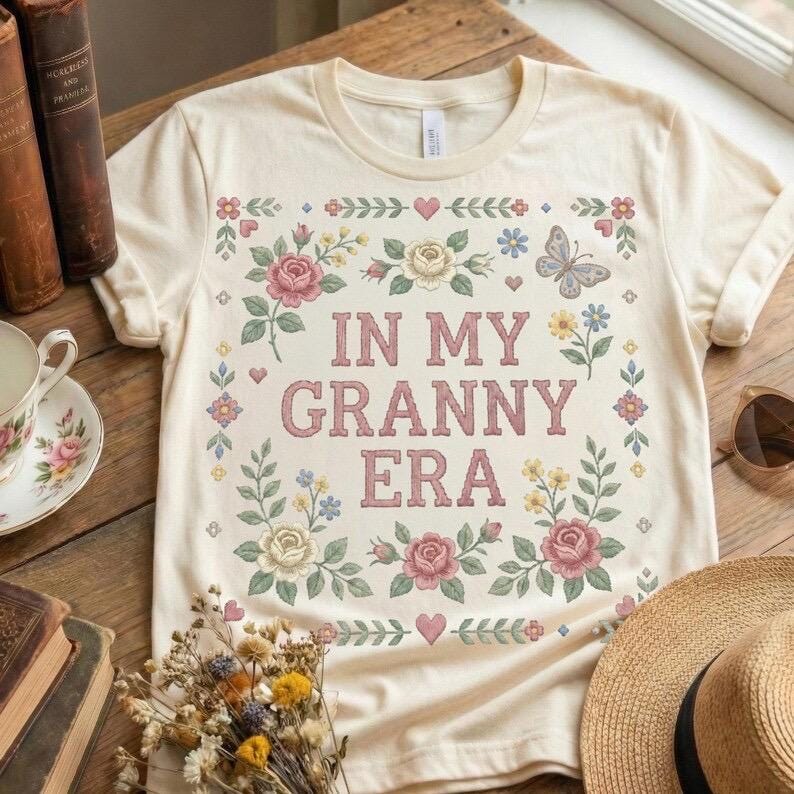 Pode incluir: Camiseta creme com o texto rosa "IN MY GRANNY ERA", rodeada por um design floral com rosas cor-de-rosa, flores azuis e uma borboleta. A camiseta est&aacute; em uma superf&iacute;cie de madeira.