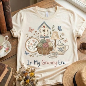 Pode incluir: Camiseta creme com um design extravagante com uma casa de p&aacute;ssaros, borboletas, uma cesta de fios e uma chaleira. O texto "In My Granny Era" &eacute; impresso em uma fonte de script. A camisa tem uma gola redonda cl&aacute;ssica.