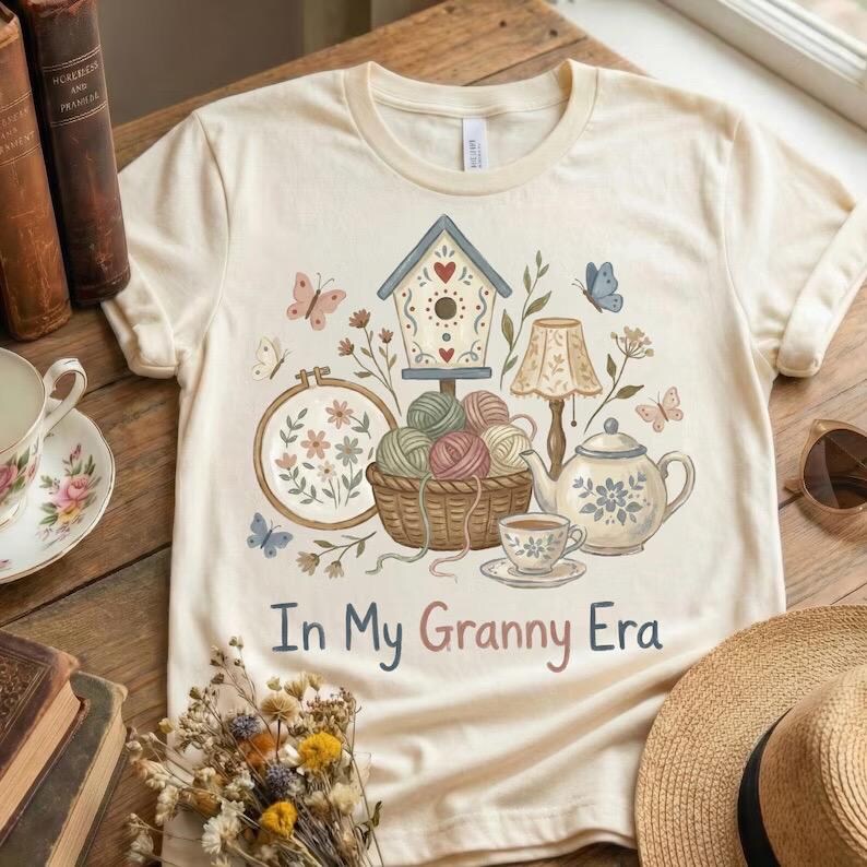 Pode incluir: Camiseta creme com um design extravagante com uma casa de p&aacute;ssaros, borboletas, uma cesta de fios e uma chaleira. O texto "In My Granny Era" &eacute; impresso em uma fonte de script. A camisa tem uma gola redonda cl&aacute;ssica.