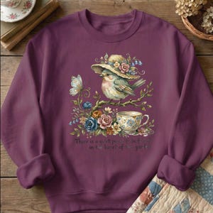 Pu&ograve; includere: Una felpa color prugna con un'illustrazione ad acquerello di un uccello con un cappello, appollaiato su un ramo con fiori e una tazza da t&egrave;. Sono inclusi anche una farfalla e il testo "There is a quiet place to be found in the heart of the garden".
