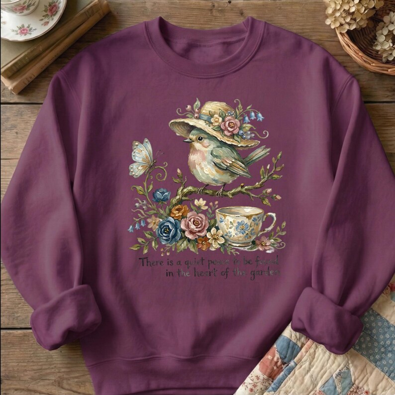 Pu&ograve; includere: Una felpa color prugna con un'illustrazione ad acquerello di un uccello con un cappello, appollaiato su un ramo con fiori e una tazza da t&egrave;. Sono inclusi anche una farfalla e il testo "There is a quiet place to be found in the heart of the garden".