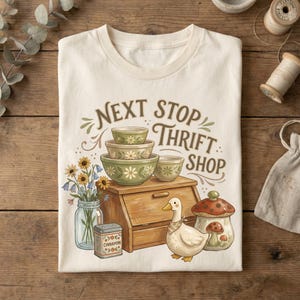 May include: Cream-colored t-shirt with a graphic featuring the text "Next Stop Thrift Shop." The design includes green bowls, a wooden breadbox, a duck, a mushroom, and a jar of flowers. A vintage-inspired design.