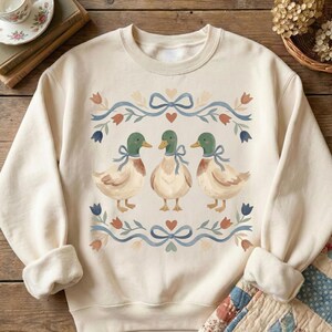May include: A cream-coloured sweatshirt with a watercolour design of three ducks, each with green heads and blue bows. The ducks are framed by a floral and ribbon border with hearts and tulips. The sweatshirt has a crew neck and long sleeves.