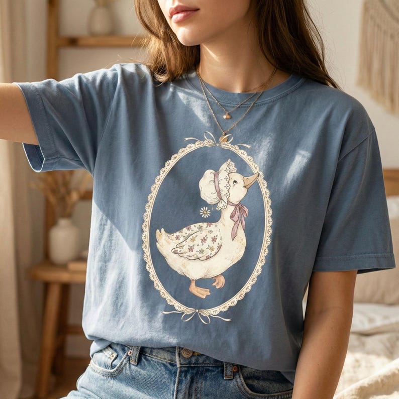 May include: A light blue t-shirt featuring a vintage-style illustration of a goose wearing a bonnet and a pink bow. The goose is inside an oval frame with a lace-like border. The shirt is made of a soft material.