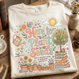 May include: Cream-colored t-shirt with a colorful garden-themed graphic. The design includes the text "Here to Plant Seeds" with flowers, a watering can, a sun, and a tree with books. The text "Let us not grow weary in doing good. Galatians 6:9" is also included.