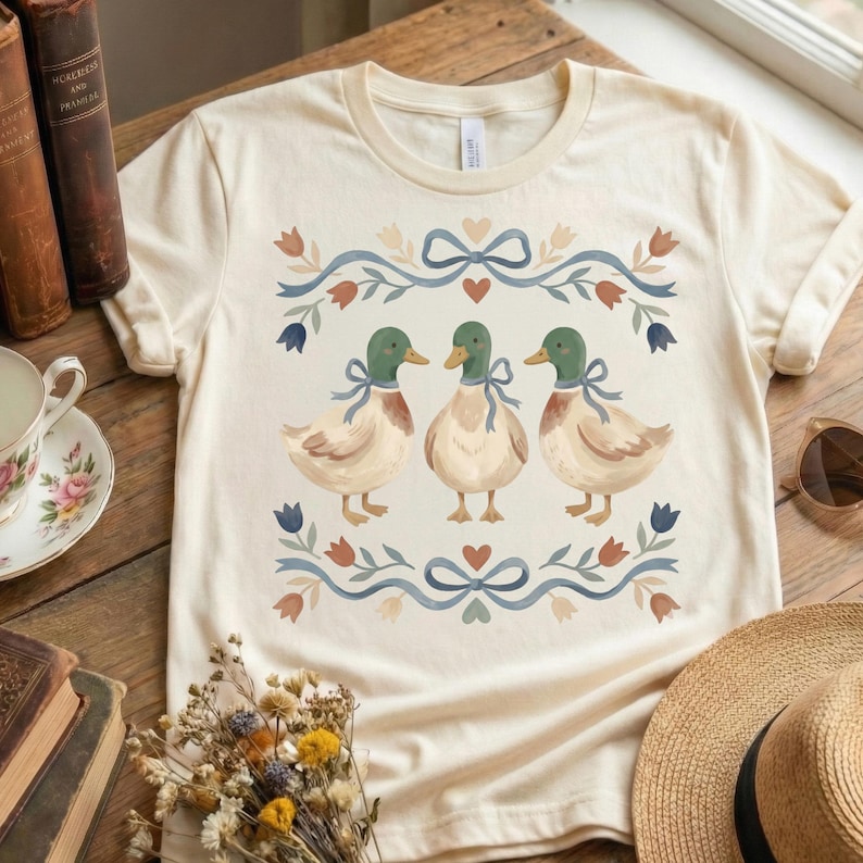 May include: A cream-coloured t-shirt with a charming illustration of three ducks, each with green heads and blue bows. The design is framed by a floral and ribbon border in blue, red, and brown. The shirt is displayed on a wooden surface.
