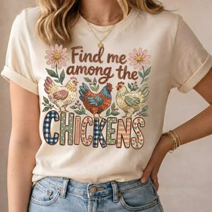May include: Cream-colored t-shirt featuring a whimsical design with three chickens and floral accents. The text "Find me among the Chickens" is displayed in a decorative font, making it a charming addition to any wardrobe.