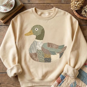 Może przedstawiać: Kremowa bluza z aplikacją w kształcie kaczki. Kaczka ma zielono-brązową kratkowaną głowę, żółty dziób i korpus z różnych wzorów tkanin w odcieniach niebieskiego, zielonego i brązowego. Bluza ma okrągły dekolt i prążkowane mankiety.