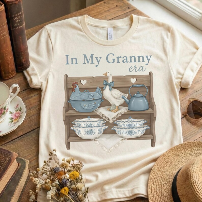 Op de afbeelding: Cr&egrave;mekleurig T-shirt met de tekst "In My Granny era" in lichtblauw. De afbeelding toont een houten plank met een blauwe kipvormige schaal, een gans met een blauwe strik en blauwe serveerschalen.